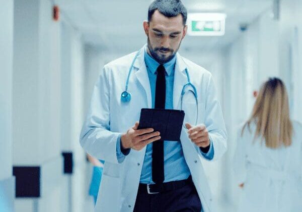 A doctor in a white coat walking down a hallway using tablet.