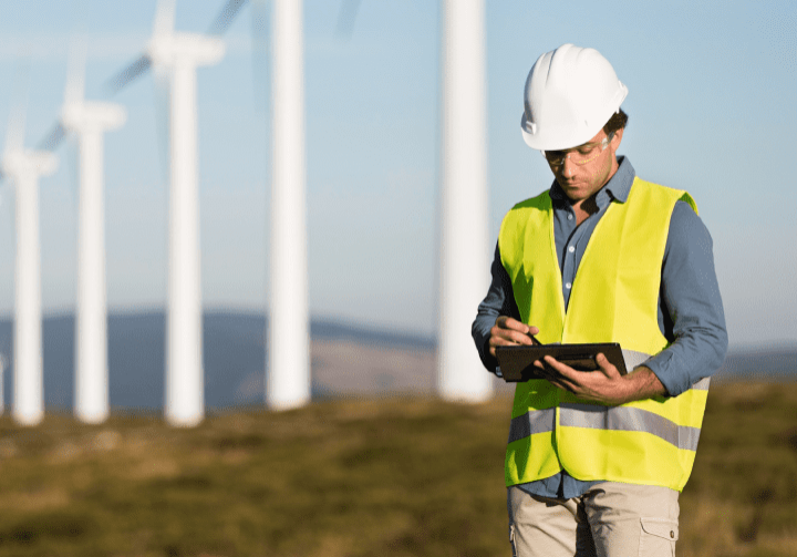 A man wearing a hard hat and vest stands in front of wind turbines, ready to work on them.